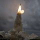 An Ariane 5 rocket shoots skyward, with bright orange fuel burned behind it, against a grey sky.